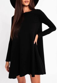 Femme portant une robe noire ample à manches longues et un chapeau noir à large bord, avec une main sur la hanche, sur un fond blanc.