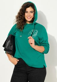 Felpa verde con cappuccio regolabile, caratterizzata da un design in bianco su sfondo nero sul petto. Abbinata a pantaloni neri texture e una borsa nera.