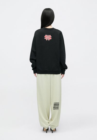 Melna džemperis ar sarkanu logo uz muguras, kombinēts ar gaiši zaļām platajām biksēm ar grafisku rakstu. Gluda auduma, brīvs griezums.