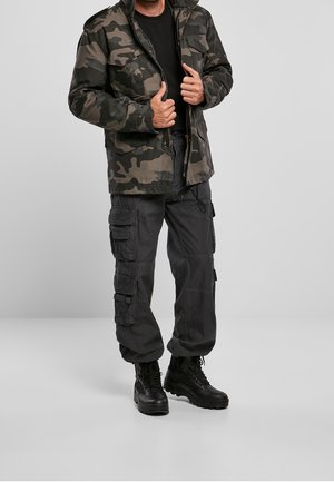 Camouflage jack in tinten van groen en bruin, gecombineerd met zwarte cargobroeken; zwarte laarzen met robuust ontwerp; eenvoudige, functionele details.