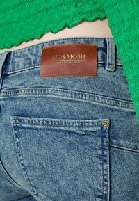 Jeans de denim azul claro con una etiqueta de cuero que dice "MOS Mosh." Presentan costuras visibles, bolsillos traseros y una textura ligeramente desgastada.