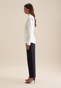 Witte blouse met lange mouwen en hoge kraag, gecombineerd met marineblauwe rechte broek en zwarte open teenhakken, zijaanzicht.