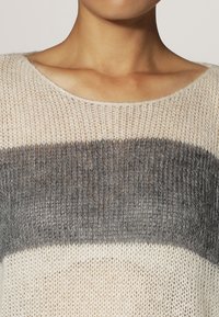 Stickad tröja med bred halsringning. Har ljus beige och grå horisontella ränder. Mjuk textur med en lös vävdesign.