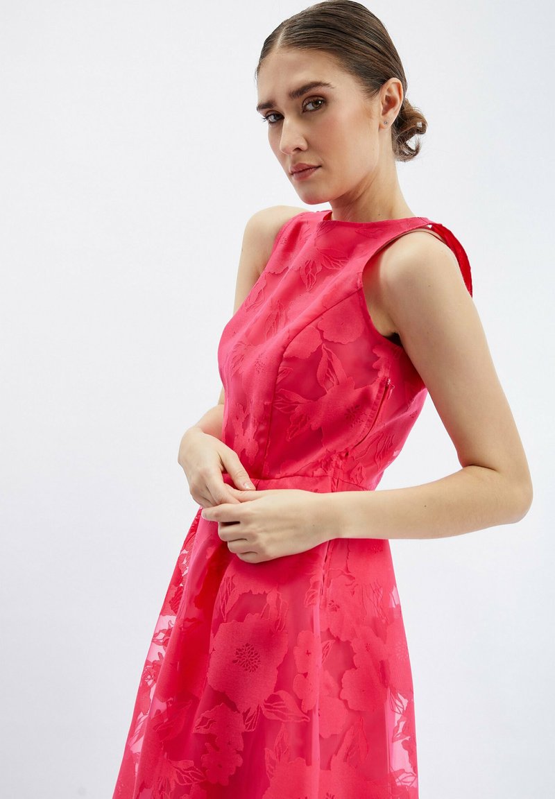 Frau mit dunklem Haar in einem tiefen Dutt, die ein ärmelloses, leuchtend pinkes Blumenkleid trägt und vor einem schlichten weißen Hintergrund posiert.