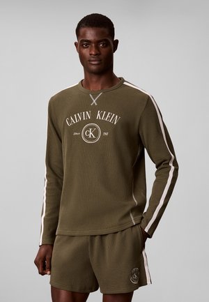 Męski model noszący oliwkowo-zielony longsleeve Calvin Klein i pasujące szorty z białymi paskami na rękawach i bokach.
