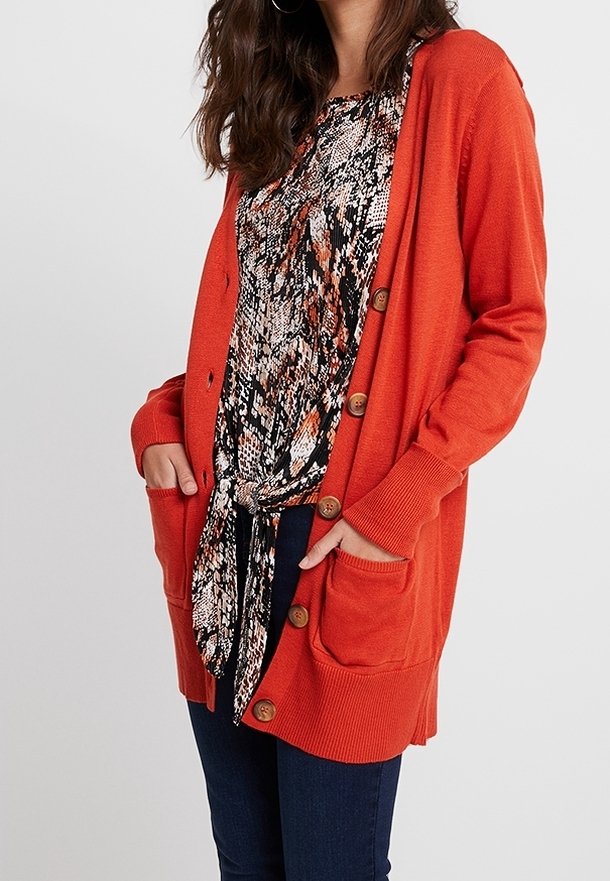 Long gilet orange avec boutons et poches avant, superposé sur un haut à motifs noirs et oranges. Présente des poignet et un ourlet côtelés.