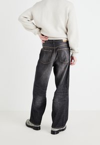 Jeans de pierna ancha negros con un lavado desgastado, que cuentan con dos bolsillos traseros y costuras naranjas, combinados con un suéter de color claro.