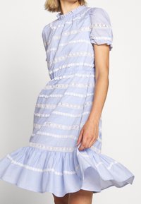 Femme en robe bleu clair avec des rayures en dentelle blanche, ourlet à volants, manches courtes bouffantes et taille froncée, tenant légèrement la robe.