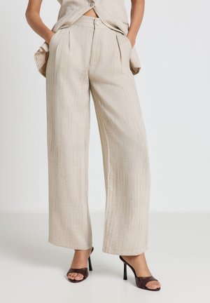 Woman wearing beige wide-leg pants with front pleats and brown high-heeled open-toe sandals, standing with hands in pockets.