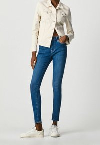 Light beige denim jacket with two chest pockets, paired with blue skinny jeans. White sneakers complete the outfit. Simple and minimal design.