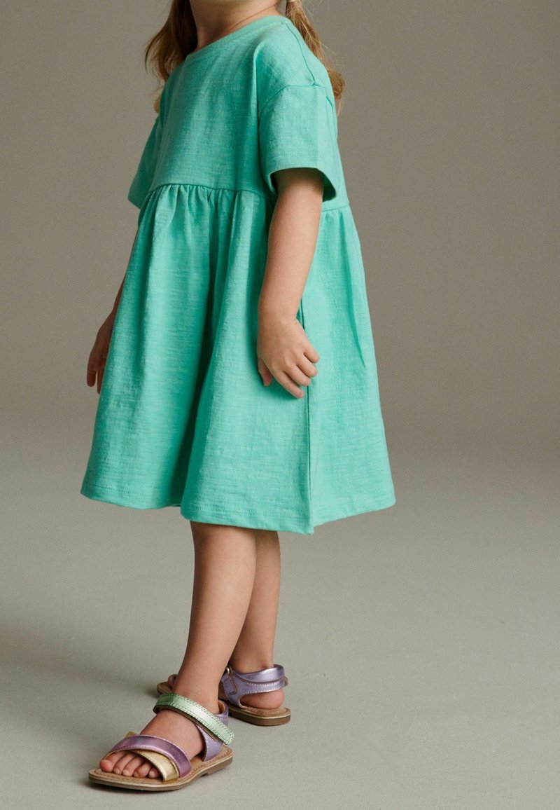 Jeune enfant portant une robe turquoise clair à manches courtes et des sandales métalliques colorées, debout devant un fond neutre.