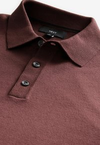 Polo de punto burdeos con un cuello acanalado, tres botones oscuros y una textura suave. La etiqueta muestra "NEXT" en una etiqueta negra.
