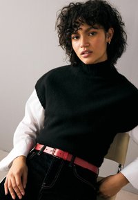 Pull en tricot sans manches noir avec un col montant, superposé sur une chemise blanche à manches longues, associé à un jean foncé et une ceinture en cuir rouge.