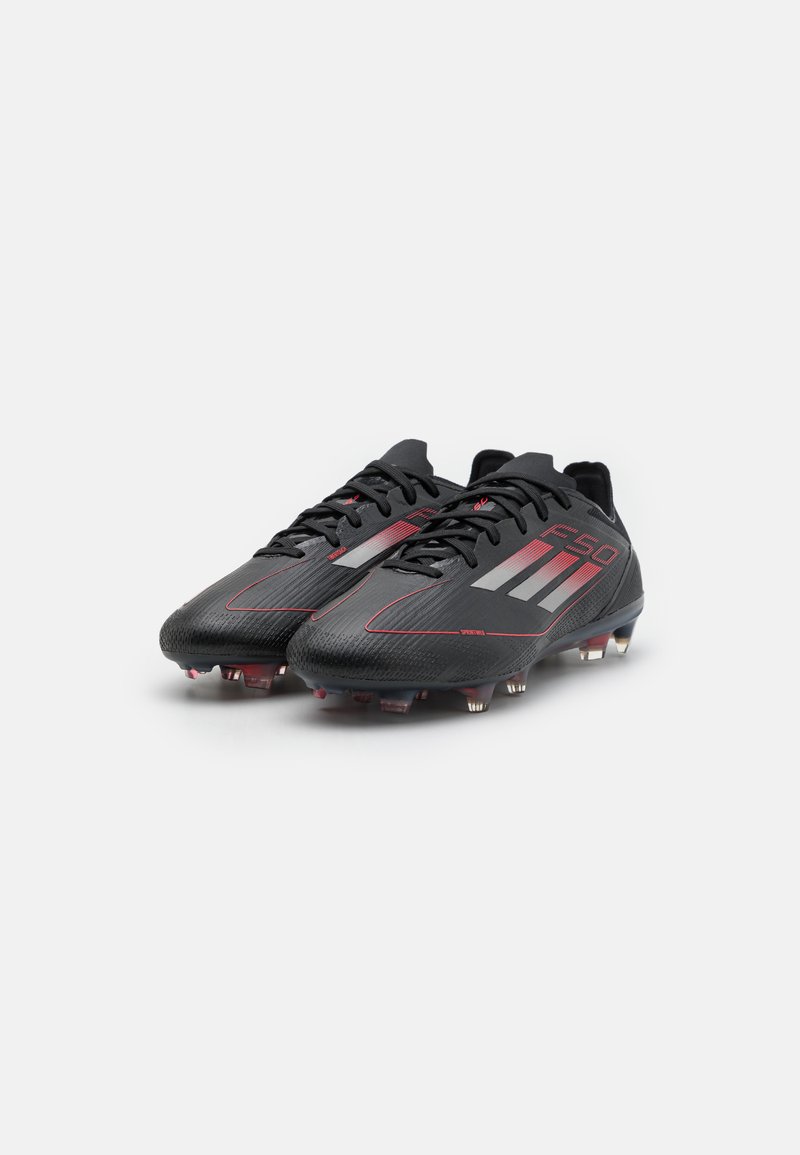 Botas de fútbol negras con acentos rojos, que presentan una superficie texturizada, cierre de cordones y clavos desmontables. El diseño incluye tres rayas blancas.