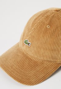 Cappello da baseball in corduroy color beige con piccolo logo di coccodrillo verde ricamato sul davanti, posizionato su uno sfondo bianco.