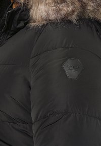 Zwarte gewatteerde winterjas met een capuchon gevoerd met bont en een zeshoekige leren patch met het label "ONLY" op de mouw.