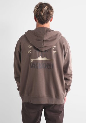 Hoodie marron avec un grand imprimé graphique représentant des palmiers et une silhouette de montagne au dos. Texture douce, coupe décontractée et capuche avec cordon de serrage.