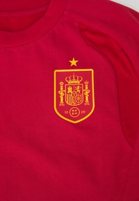Κόκκινη αθλητική φανέλα με στρογγυλή λαιμόκοψη, διακοσμημένη με ένα κίτρινο έμβλημα σε σχήμα ασπίδας που έχει στέμμα και περίπλοκα σχέδια στο αριστερό στήθος.