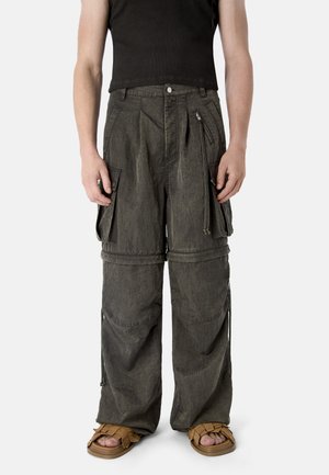 Osoba ubrana w czarny bezrękawnik, ciemnoszare szerokie spodnie cargo z wieloma kieszeniami i zamkami błyskawicznymi oraz jasnobrązowe plecione sandały.