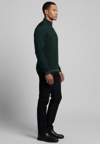 Jersey verde de canalé con cuello alto y detalles negros en los hombros. Combinado con pantalones y botas negras, visto de perfil.