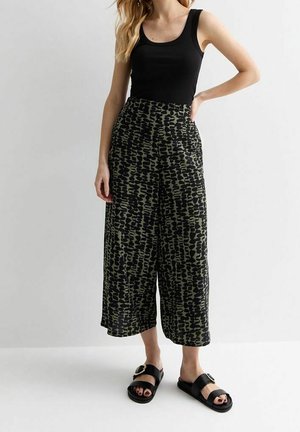 Femme portant un haut noir sans manches, un pantalon taille haute à jambes larges avec un motif abstrait noir et olive, et des sandales noires à boucle.