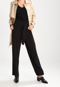 Abrigo trench beige sobre una blusa negra de escote en v, combinado con pantalones negros de pierna ancha, ceñidos a la cintura, y mules negros de tacón grueso.