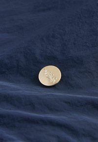 Pulsante rotondo in metallo con finitura oro spazzolato, caratterizzato da un design floreale in rilievo, appoggiato su un tessuto navy stropicciato.