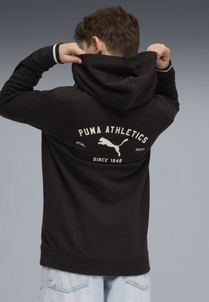 Persona joven ajustándose la capucha de una sudadera negra de Puma Athletics con logo y texto en la parte trasera, vistiendo jeans azul claro.
