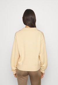 Beige, stickad tröja med avslappnad passform, stor krage och ribbat manschettdetalj. Mjuk textur, når ner till höfterna, i kombination med bruna veckade byxor.