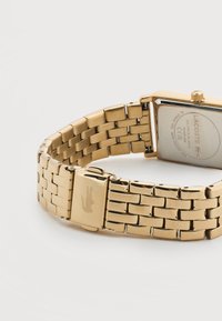 Ρολόι βραχιόλι από μέταλλο σε χρυσό τόνο με ορθογώνια θήκη, λογότυπο Lacoste στην αγκράφα και χαραγμένες λεπτομέρειες της μάρκας στην πίσω όψη.