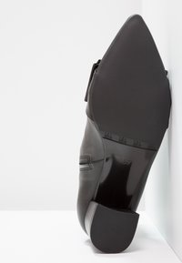 Zapato de vestir de cuero negro reposando con la suela hacia arriba contra una pared blanca, con la suela lisa mostrando la talla 37 grabada cerca del talón.