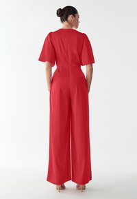 Red jumpsuit with short, wide sleeves, a fitted waist, and wide-leg trousers. Smooth texture and seamless back design, complemented by a zipper.