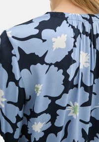 Blauwe bloemetjesblouse met grote marineblauwe en lichtblauwe bloemen, aangevuld met witte en groene accenten. Soepele textuur met geplooid achterpand.