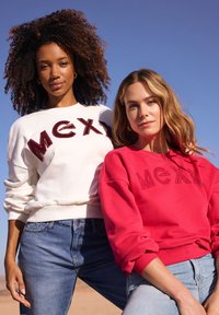 Dos mujeres llevan sudaderas con la palabra "MEXI" en letras gruesas y suaves; una en blanco y otra en rojo. Ambas combinadas con jeans de mezclilla. Fondo de cielo azul claro.