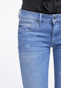 Ljusblå denimjeans med en åtsittande design, som har en framficka, synlig sömnad, subtila slitningar och en klassisk knappstängning.