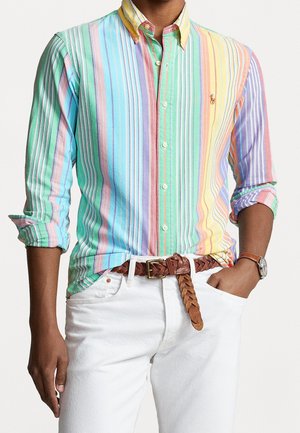 Homme portant une chemise à rayures verticales multicolores boutonnée avec les manches retroussées, un pantalon blanc, une ceinture tressée marron et une montre-bracelet.