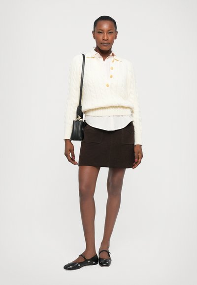 Pull en tricot blanc à cols ronds et boutons, superposé à une chemise claire. Jupe mini en daim marron avec poches et ballerines noires.