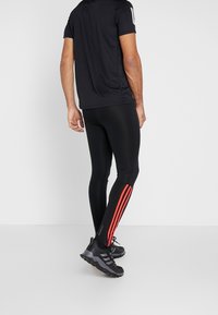 Haut de sport noir à manches courtes, associé à un legging noir avec des rayures latérales rouges et des chaussures de sport noires avec des semelles texturées.