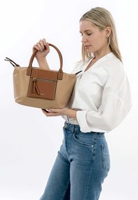 Beige tote bag dengan aksen kulit coklat, saku resleting depan, dan pegangan ganda. Permukaan halus dengan detail jahitan kontras.