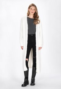 Dlouhý bílý nadýchaný kardigan s otevřeným předním dílem, nošený přes šedý top a černé roztrhané skinny džíny. Černé vysoké kozačky dotvářejí outfit.