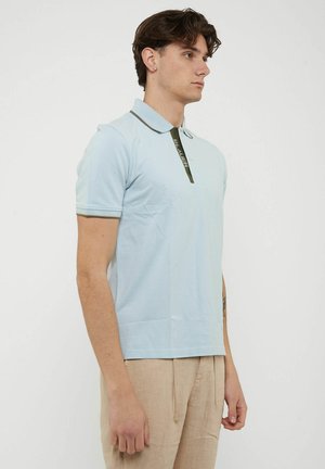 Giovane uomo con polo azzurro chiaro con rifiniture verde scuro e pantaloni beige, in piedi di lato su sfondo bianco uniforme.