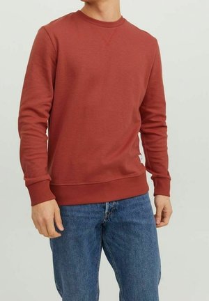 Mies, jolla on ruosteenvärinen pyöreäkauluksinen collegepaita ja siniset farkut, seisomassa rentoina käsivarsina yksinkertaisen vaalean taustan edessä.