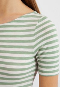 Kurzärmliges Oberteil mit grünen und weißen Streifen, das über einen weiten Halsausschnitt und einen weichen, strukturierten Stoff verfügt. Betont mit eng anliegenden Ärmeln.