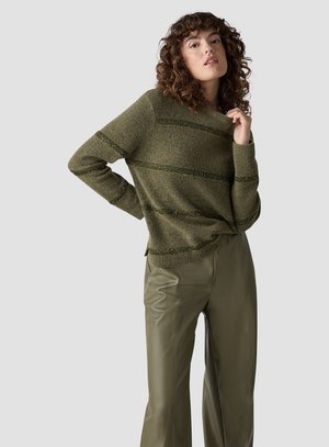 Frau mit lockigem Haar, die einen olivgrün gestreiften Pullover und passende weit geschnittene Lederhosen trägt und mit einer Hand nahe am Gesicht posiert.