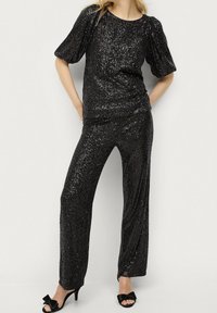Top à sequins noirs et pantalon assorti, avec une coupe décontractée et des manches bouffantes. Mis en valeur par une texture brillante et un léger éclat. Escarpins noirs.