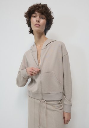 Junge Person mit kurzen lockigen Haaren, die einen beigen Reißverschluss-Hoodie und passende Hosen trägt, steht vor einem einfarbigen hellen Hintergrund.