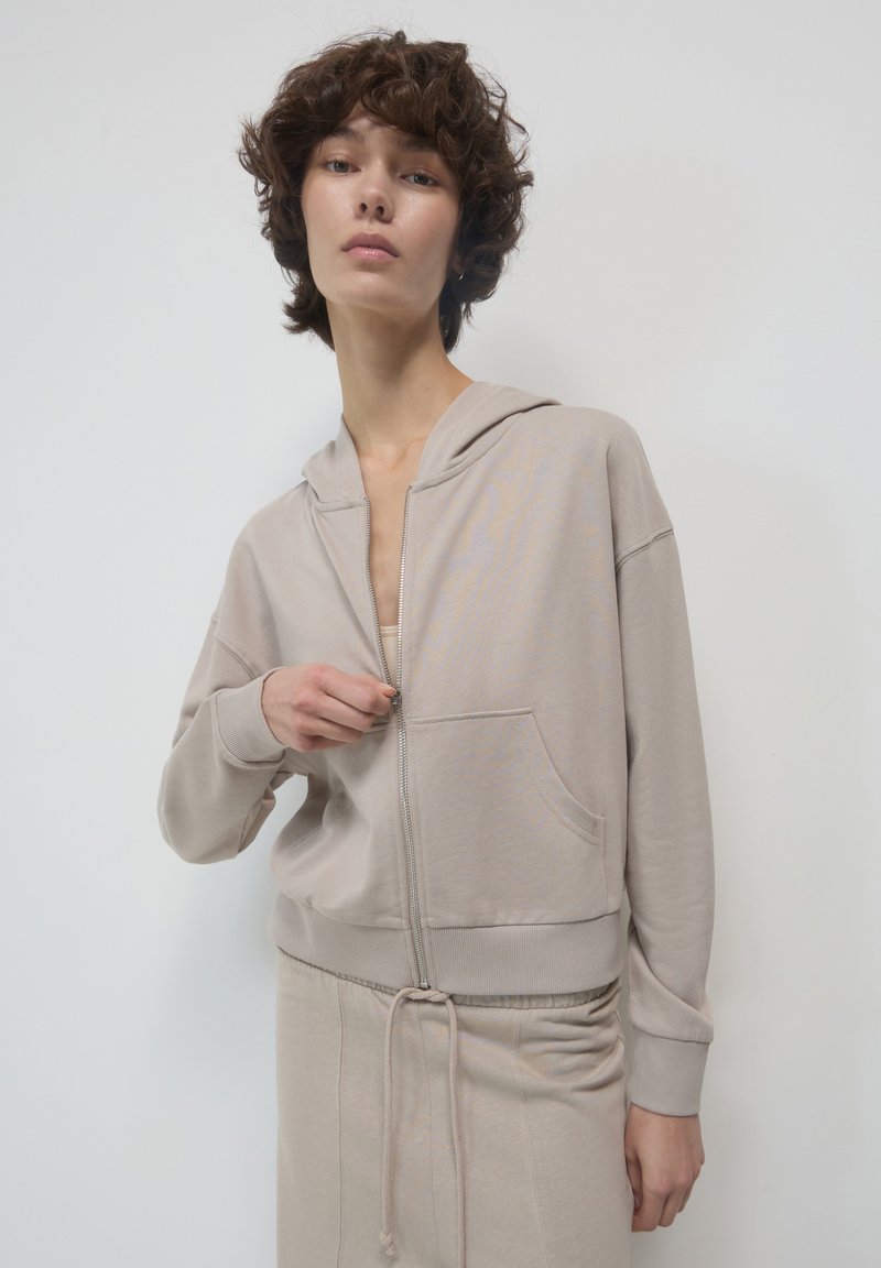 Giovane con capelli corti e ricci, che indossa una felpa beige con zip e pantaloni coordinati, in piedi contro uno sfondo chiaro e semplice.