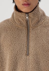 Beige fleece trui met hoge kraag, voorzien van een metalen ritssluiting bij de hals. De stof heeft een gestructureerde, zachte uitstraling.
