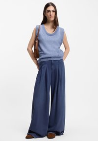 Hellblaues, ärmelloses Strickoberteil mit rundem Halsausschnitt, kombiniert mit weit geschnittenen Marineblauen Hosen. Eine braune Umhängetasche liegt auf der linken Seite.