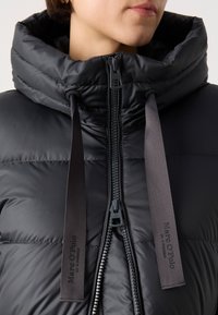 Chaqueta de plumas negra con un alto cuello, cierre con cremallera y dos cordones de tela negra con el logotipo de la marca. Textura de superficie suave y brillante.